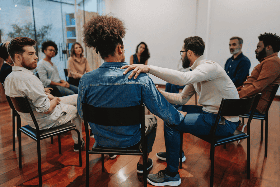 Support group seated in a circle, with one participant offering reassurance to another, representing the importance of community in helping someone with alcoholism.