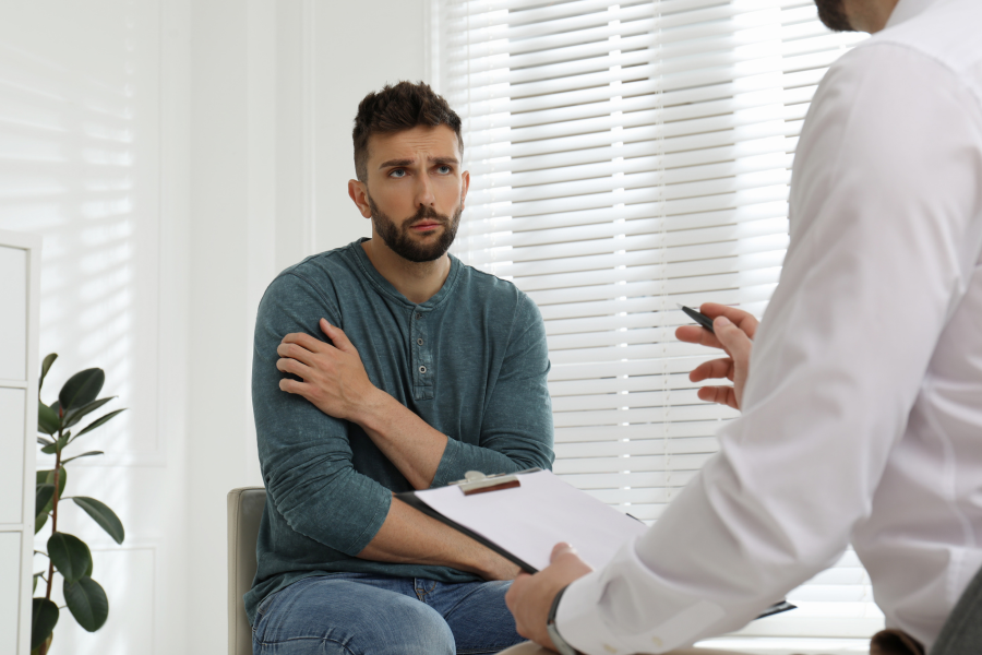 Concerned man in a teal shirt sitting in a therapist’s office, holding his shoulder while talking to a doctor with a clipboard.